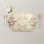 An oval embroidered table mat / placemat in cream satin with pink floral motifs and gold-toned lace trim, styled on a neutral beige surface with cutlery and white flowers.
