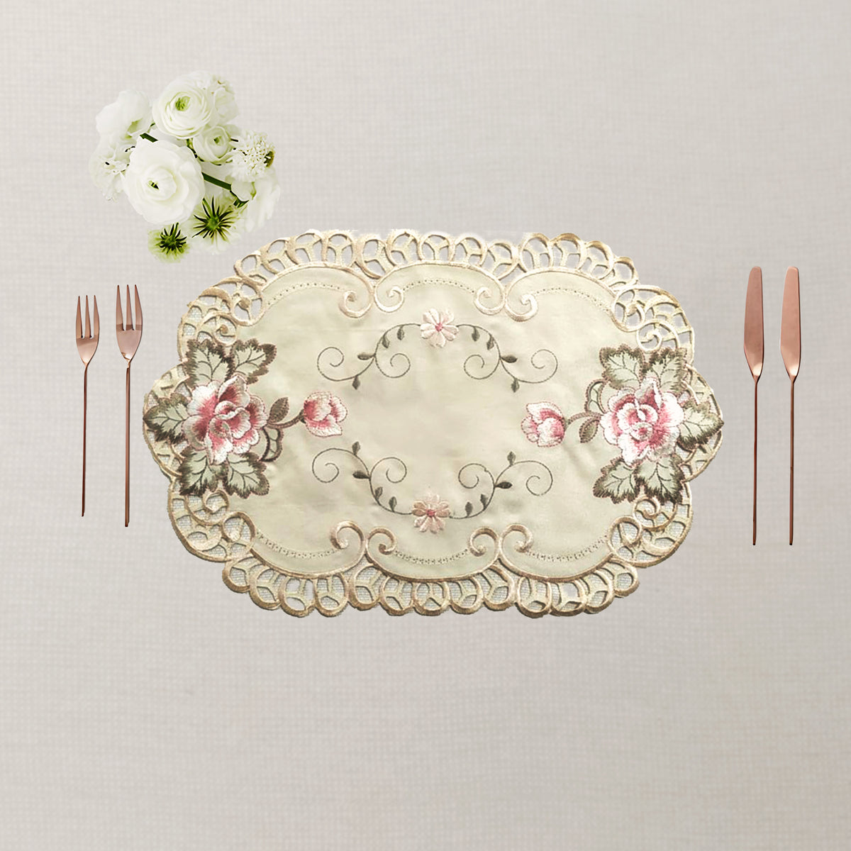 An oval embroidered table mat / placemat in cream satin with pink floral motifs and gold-toned lace trim, styled on a neutral beige surface with cutlery and white flowers.
