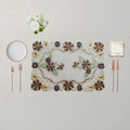 An embroidered rectangular placemat with a floral pattern in brown, beige, and green, styled on a light beige tablecloth with a white plate, two rose gold forks, two rose gold knives, a folded napkin, and a small bouquet of white flowers.