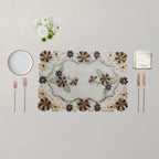 An embroidered rectangular placemat with a floral pattern in brown, beige, and green, styled on a light beige tablecloth with a white plate, two rose gold forks, two rose gold knives, a folded napkin, and a small bouquet of white flowers.