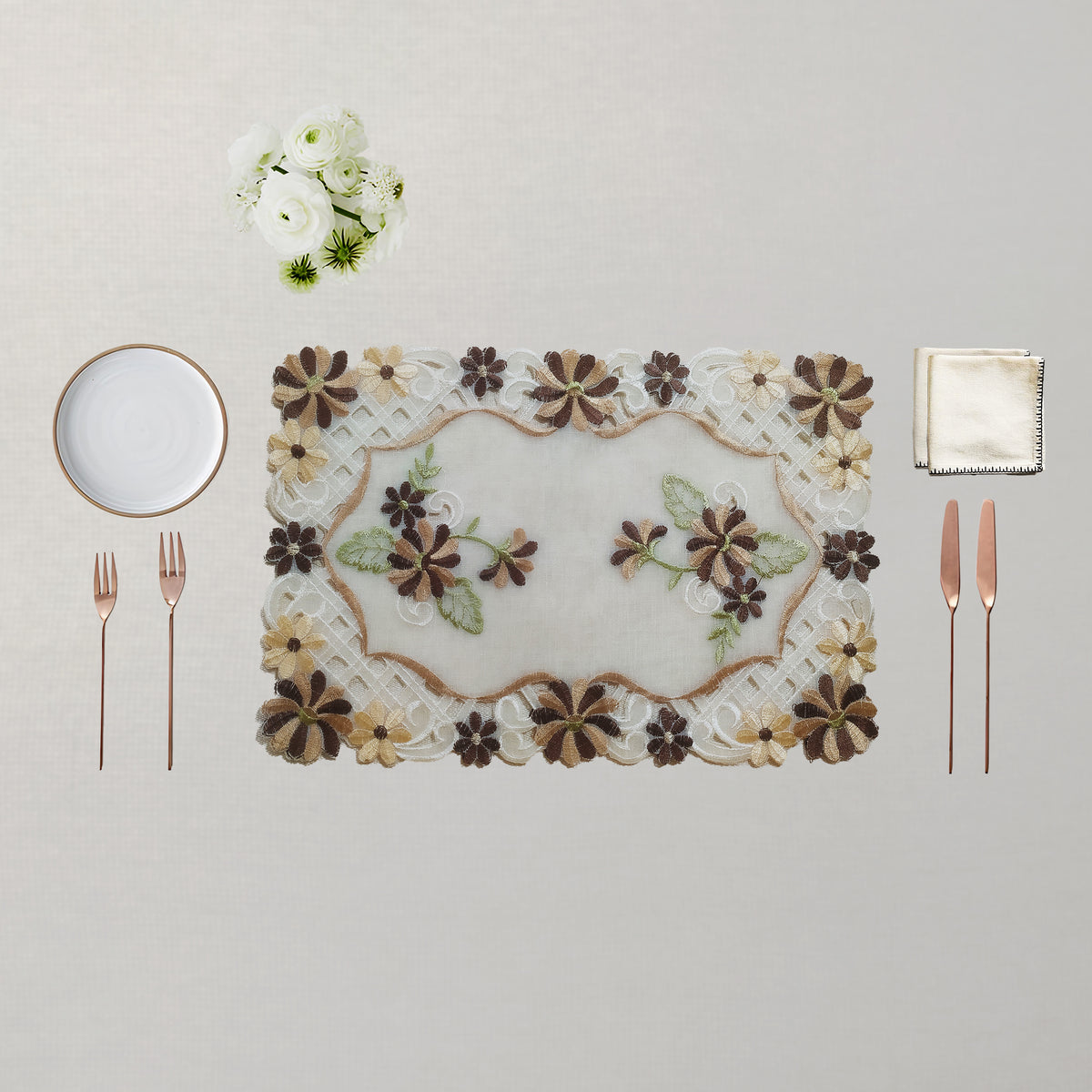 An embroidered rectangular placemat with a floral pattern in brown, beige, and green, styled on a light beige tablecloth with a white plate, two rose gold forks, two rose gold knives, a folded napkin, and a small bouquet of white flowers.