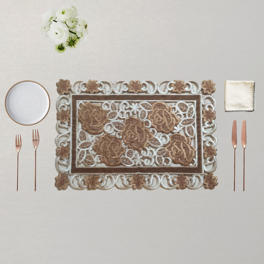 An ornate rectangular place setting featuring a decorative placemat with a raised floral pattern, accompanied by a white plate, two rose gold forks, two rose gold knives, and a white napkin on a neutral beige background.