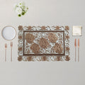An ornate rectangular place setting featuring a decorative placemat with a raised floral pattern, accompanied by a white plate, two rose gold forks, two rose gold knives, and a white napkin on a neutral beige background.