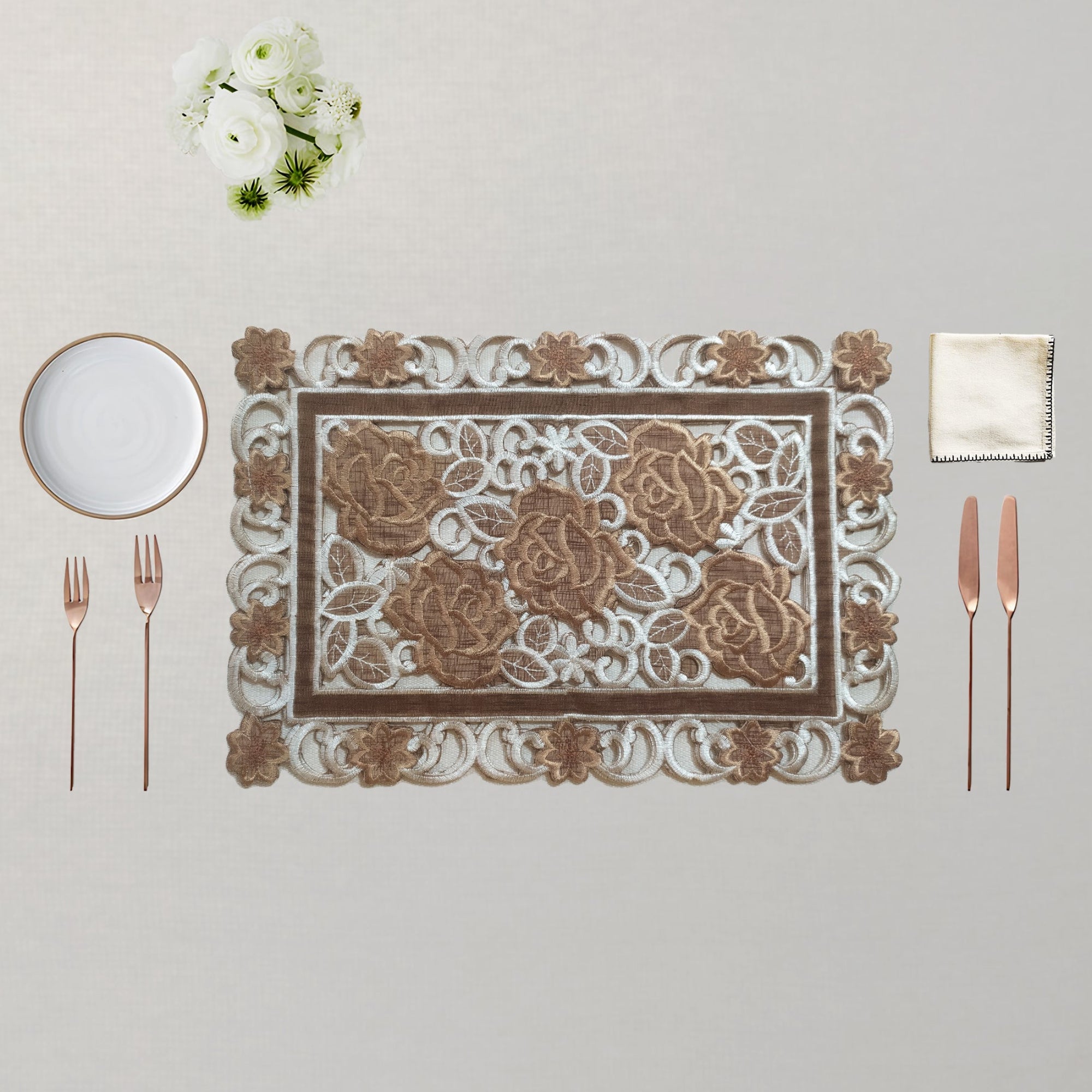 An ornate rectangular place setting featuring a decorative placemat with a raised floral pattern, accompanied by a white plate, two rose gold forks, two rose gold knives, and a white napkin on a neutral beige background.