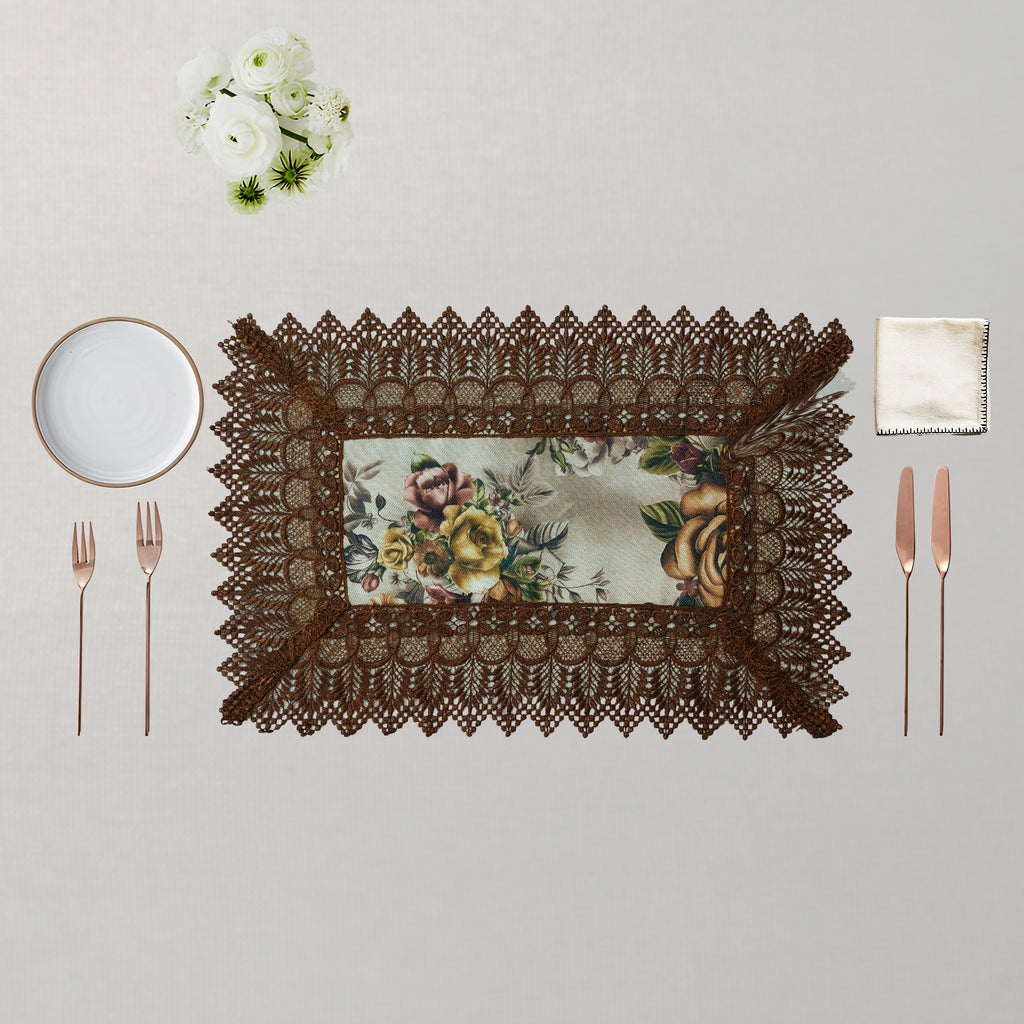A rectangular table setting featuring a dark brown lace placemat with a vintage floral pattern insert, styled on a light beige textured background with a white plate, rose gold cutlery, a cream napkin, and a small white flower bouquet.