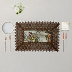 A rectangular table setting featuring a dark brown lace placemat with a vintage floral pattern insert, styled on a light beige textured background with a white plate, rose gold cutlery, a cream napkin, and a small white flower bouquet.