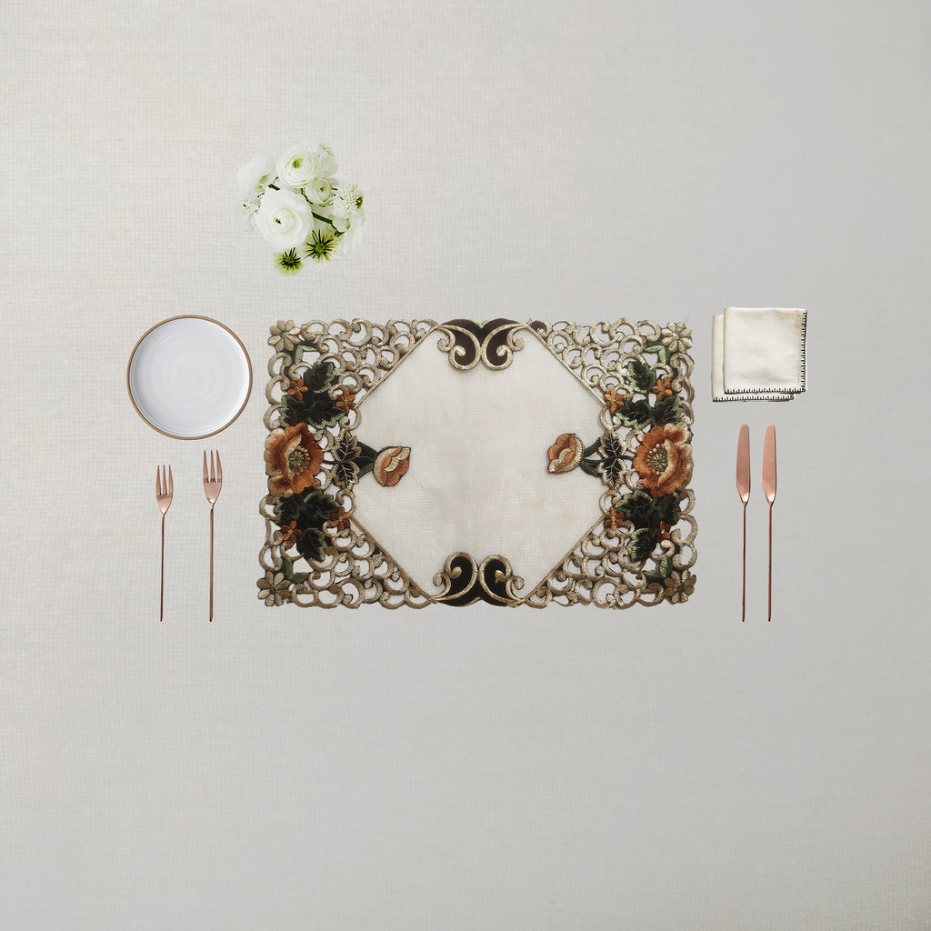 An ornate rectangular placemat with a floral and scroll pattern in beige, brown, and green, styled on a light beige tablecloth with a white plate, silver forks, rose gold cutlery, a folded napkin, and a small white flower arrangement.