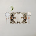 An ornate rectangular placemat with a floral and scroll pattern in beige, brown, and green, styled on a light beige tablecloth with a white plate, silver forks, rose gold cutlery, a folded napkin, and a small white flower arrangement.