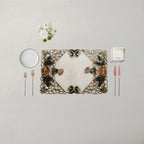 An ornate rectangular placemat with a floral and scroll pattern in beige, brown, and green, styled on a light beige tablecloth with a white plate, silver forks, rose gold cutlery, a folded napkin, and a small white flower arrangement.