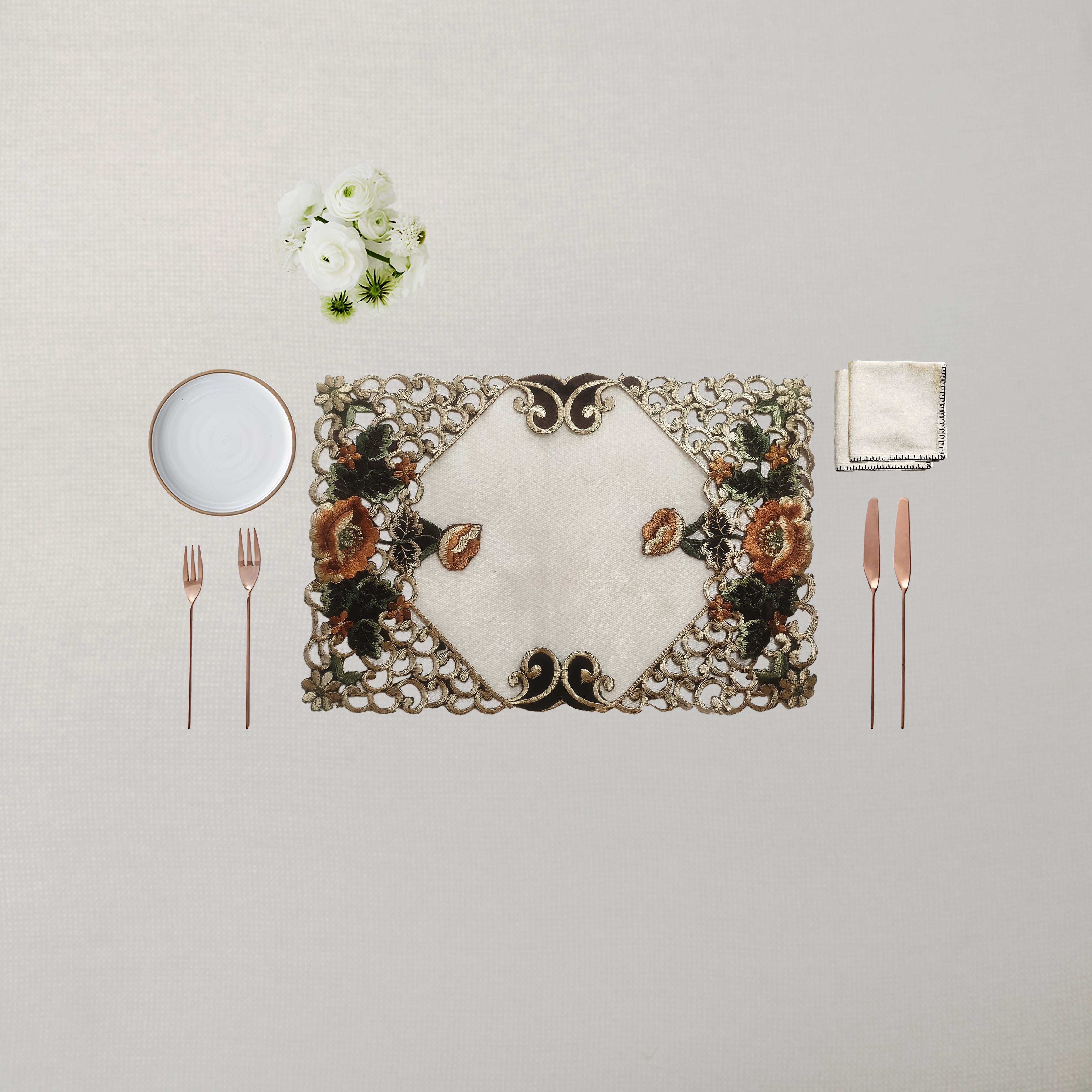 An ornate rectangular placemat with a floral and scroll pattern in beige, brown, and green, styled on a light beige tablecloth with a white plate, silver forks, rose gold cutlery, a folded napkin, and a small white flower arrangement.