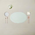 An elegant flat-lay table setting featuring a large oval white lace doily placemat in the center, accompanied by a small white plate, rose gold cutlery, a folded cream napkin, and a small bouquet of white flowers, all arranged on a textured beige linen tablecloth.