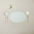 An elegant flat-lay table setting featuring a large oval white lace doily placemat in the center, accompanied by a small white plate, rose gold cutlery, a folded cream napkin, and a small bouquet of white flowers, all arranged on a textured beige linen tablecloth.