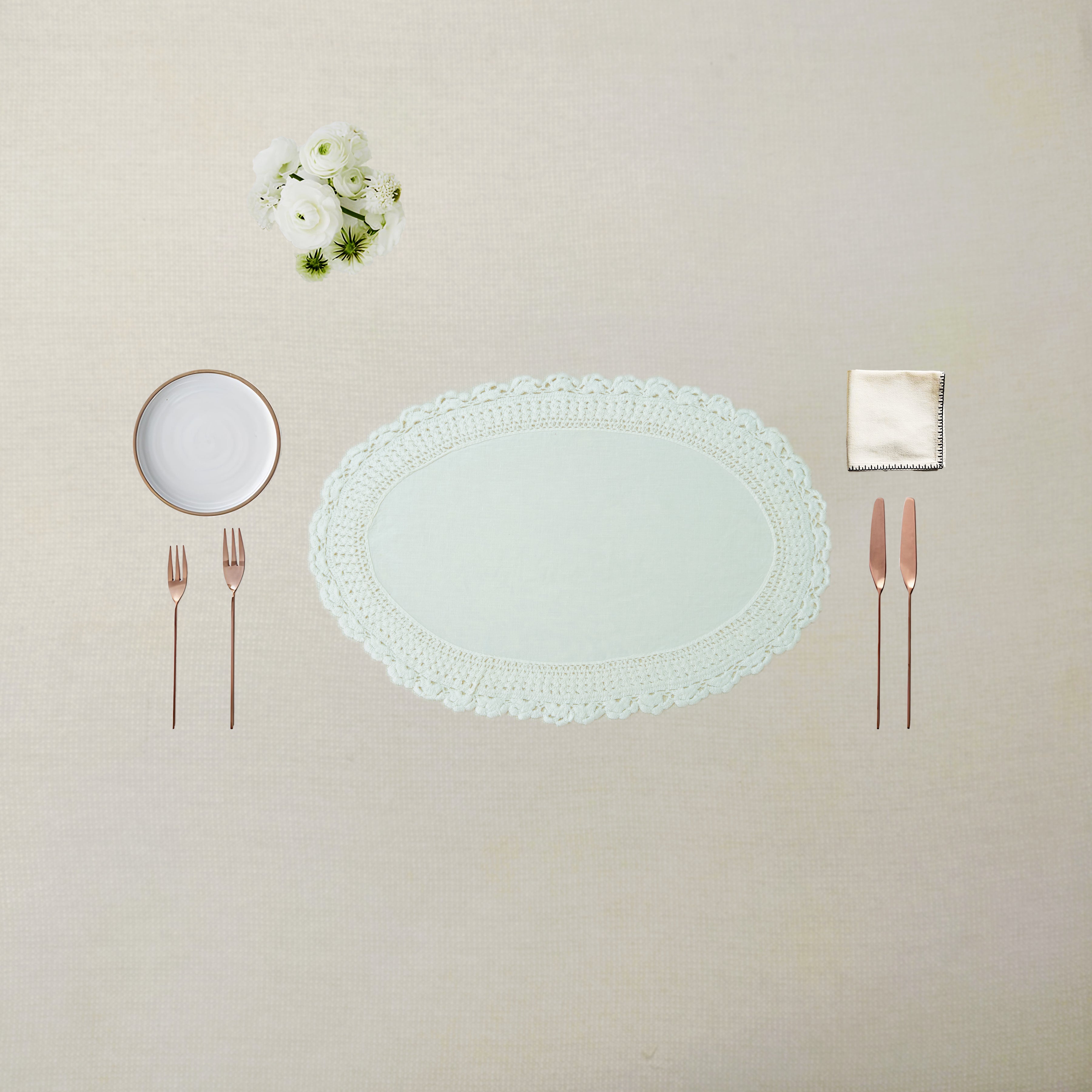 An elegant flat-lay table setting featuring a large oval white lace doily placemat in the center, accompanied by a small white plate, rose gold cutlery, a folded cream napkin, and a small bouquet of white flowers, all arranged on a textured beige linen tablecloth.