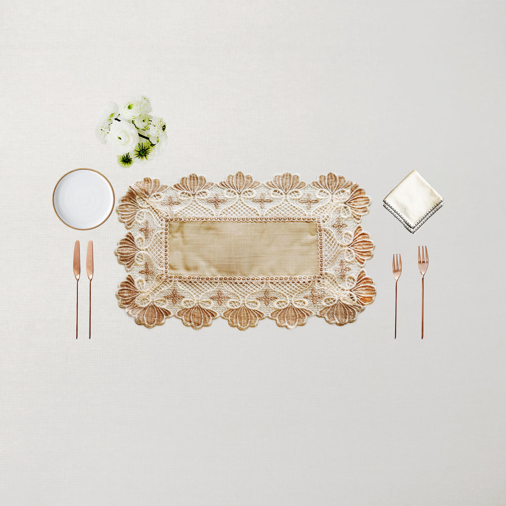 An elegant rectangular lace placemat in beige and cream with a scalloped floral border, styled in a flat-lay setting with rose gold cutlery, a white napkin, a small white plate, and a tiny bouquet of white flowers on a light beige background.