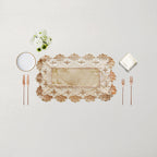 An elegant rectangular lace placemat in beige and cream with a scalloped floral border, styled in a flat-lay setting with rose gold cutlery, a white napkin, a small white plate, and a tiny bouquet of white flowers on a light beige background.