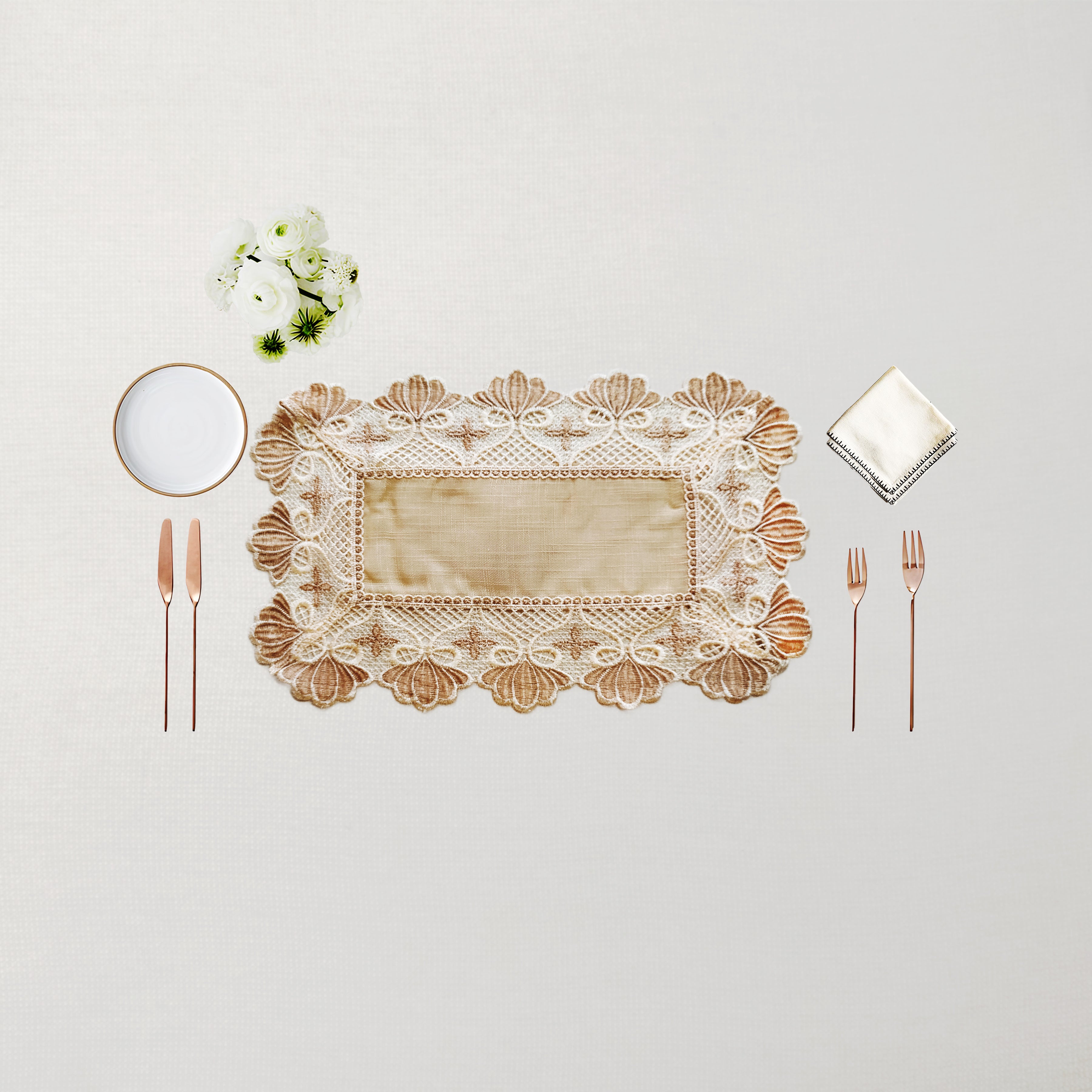 An elegant rectangular lace placemat in beige and cream with a scalloped floral border, styled in a flat-lay setting with rose gold cutlery, a white napkin, a small white plate, and a tiny bouquet of white flowers on a light beige background.