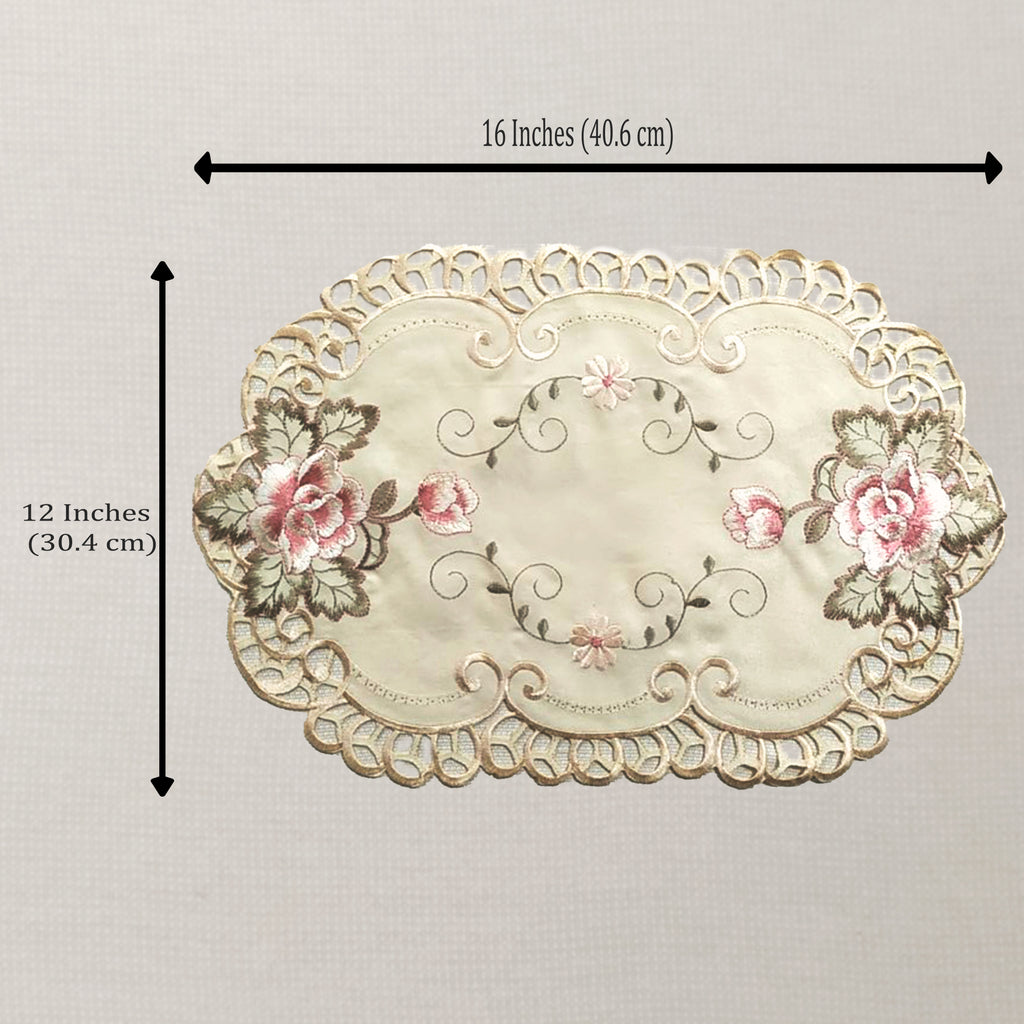 An oval embroidered placemat with a scalloped lace-like border, pink floral motifs, and gray-green leaves on a cream background. Black measurement arrows and text indicate dimensions of 16 inches by 12 inches.