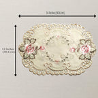 An oval embroidered placemat with a scalloped lace-like border, pink floral motifs, and gray-green leaves on a cream background. Black measurement arrows and text indicate dimensions of 16 inches by 12 inches.