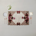 An ornate rectangular table placemat with a cream center and intricate floral embroidery in burgundy, pink, and green, styled on a neutral beige table with cutlery, a small plate, and white flowers.