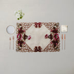 An ornate rectangular table placemat with a cream center and intricate floral embroidery in burgundy, pink, and green, styled on a neutral beige table with cutlery, a small plate, and white flowers.