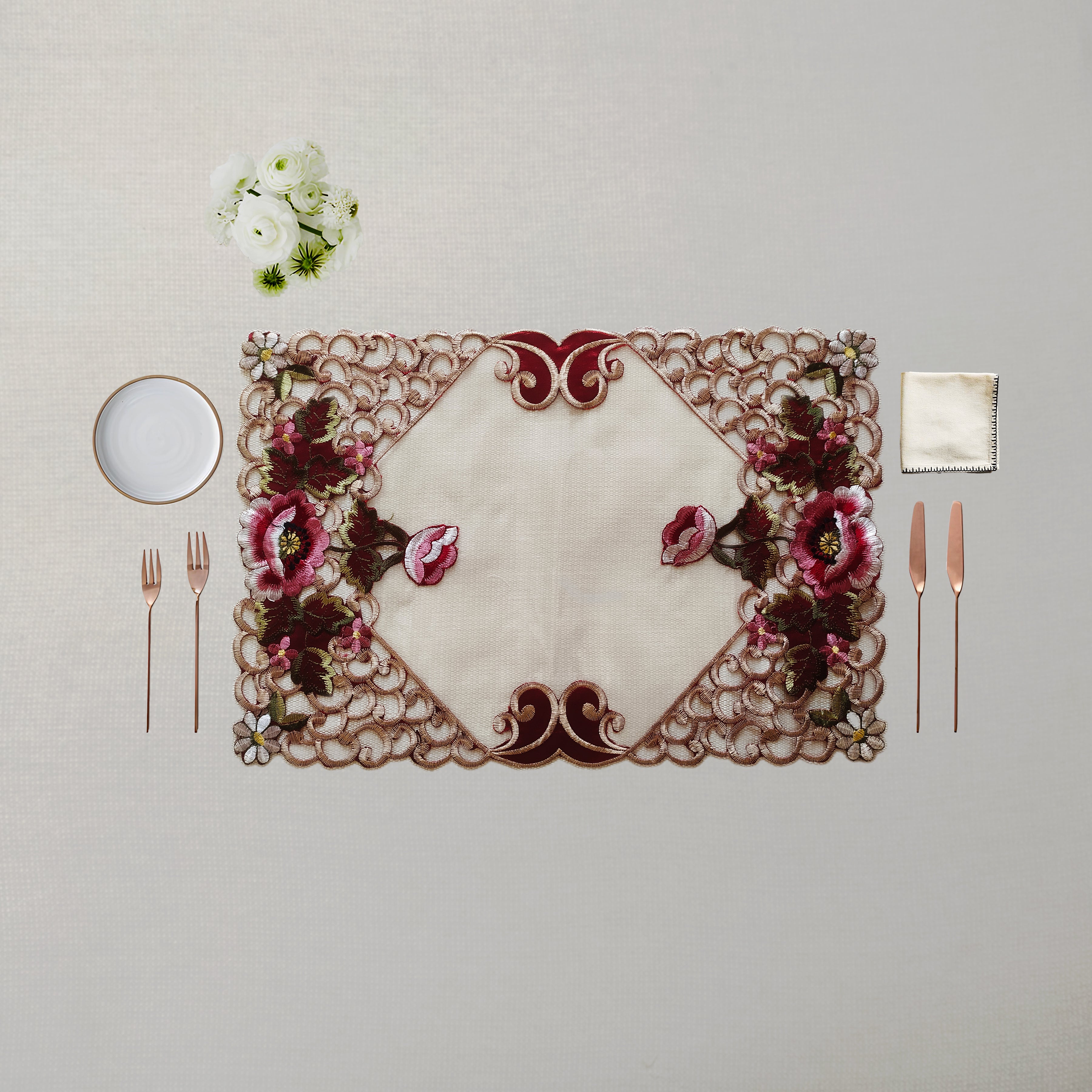 An ornate rectangular table placemat with a cream center and intricate floral embroidery in burgundy, pink, and green, styled on a neutral beige table with cutlery, a small plate, and white flowers.