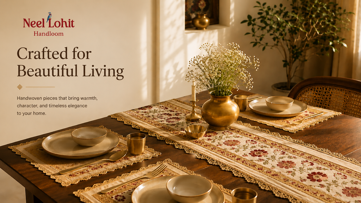 A styled dining table set with floral embroidered table runners, matching placemats, plates, bowls, and small brass vessels  and a potted plant in a warm, sunlit room. The left side features a large white panel with the text 'Neel Lohit Handloom' and 'Crafted for Beautiful Living'. This is the banner/hero image for Neel Lohit handloom hosted at neellohit.com. 