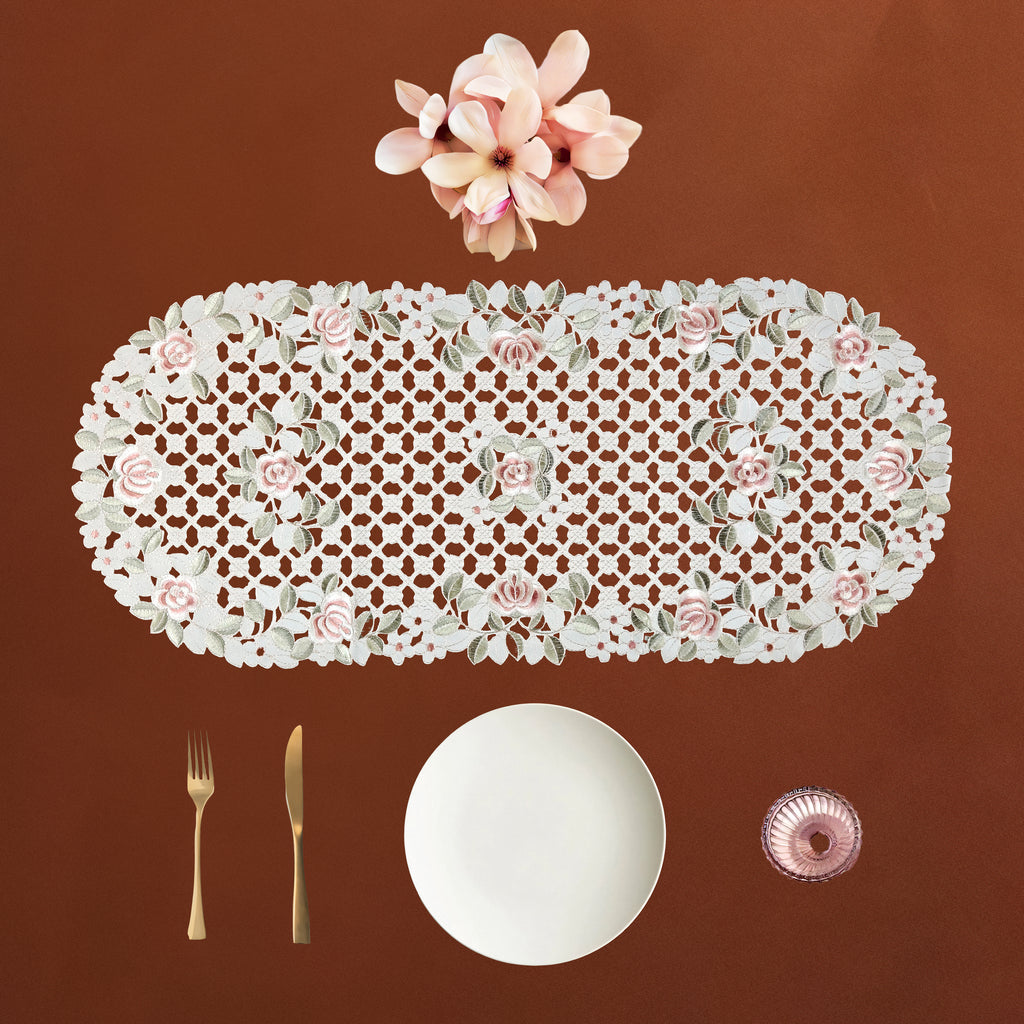An oval cream lace table runner with pink roses and green leaves, displayed on a brown background with a white plate, gold fork and knife, and a pink glass bowl.