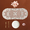An oval cream lace table runner with pink roses and green leaves, displayed on a brown background with a white plate, gold fork and knife, and a pink glass bowl.