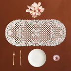 An oval cream lace table runner with pink roses and green leaves, displayed on a brown background with a white plate, gold fork and knife, and a pink glass bowl.