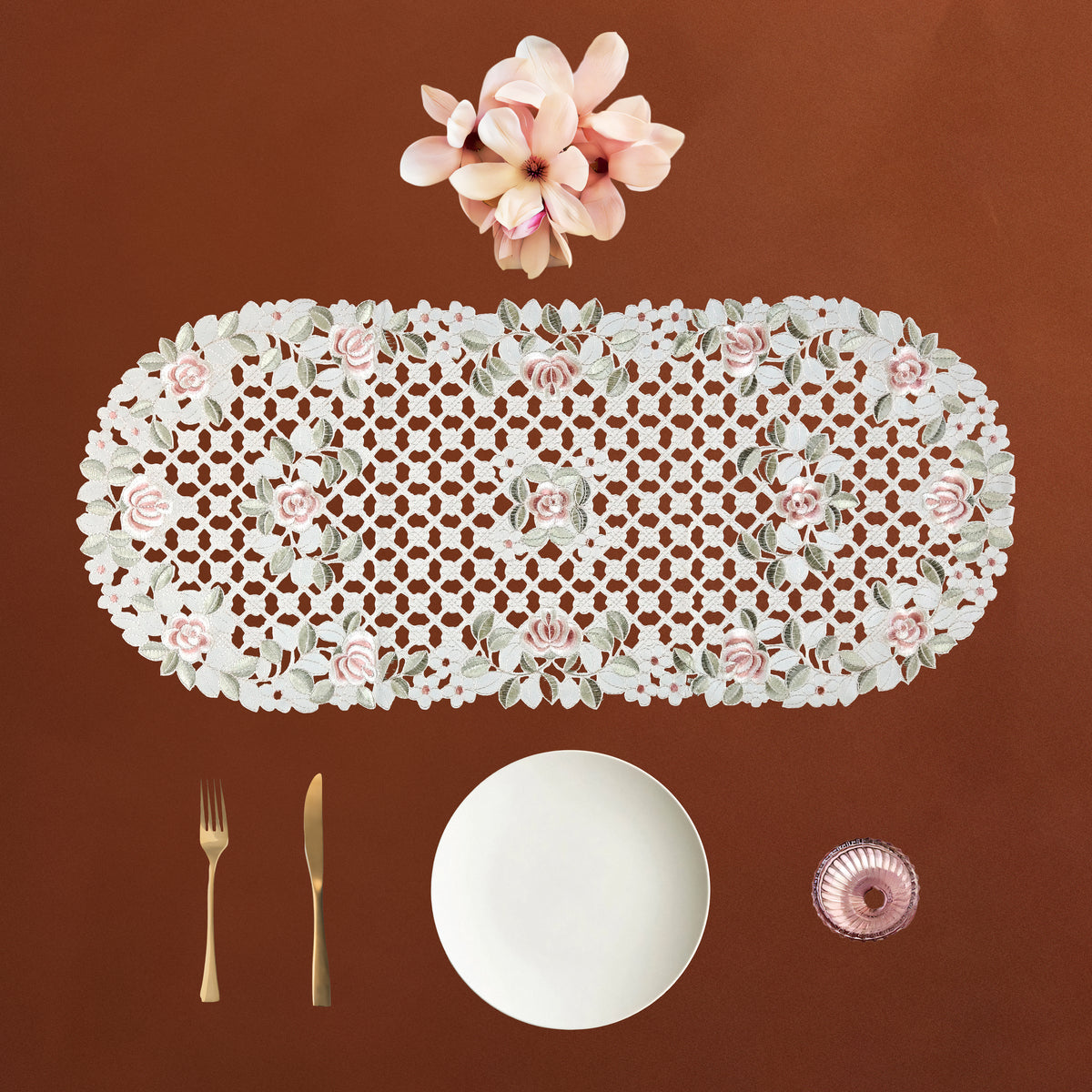 An oval cream lace table runner with pink roses and green leaves, displayed on a brown background with a white plate, gold fork and knife, and a pink glass bowl.
