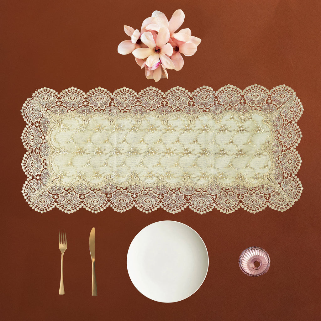 An off-white lace rectangular table runner with scalloped edges is displayed on a warm brown background, styled with a white plate, gold-toned cutlery, a small pink bowl, and pink flowers above it.