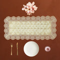 An off-white lace rectangular table runner with scalloped edges is displayed on a warm brown background, styled with a white plate, gold-toned cutlery, a small pink bowl, and pink flowers above it.
