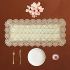 An off-white lace rectangular table runner with scalloped edges is displayed on a warm brown background, styled with a white plate, gold-toned cutlery, a small pink bowl, and pink flowers above it.