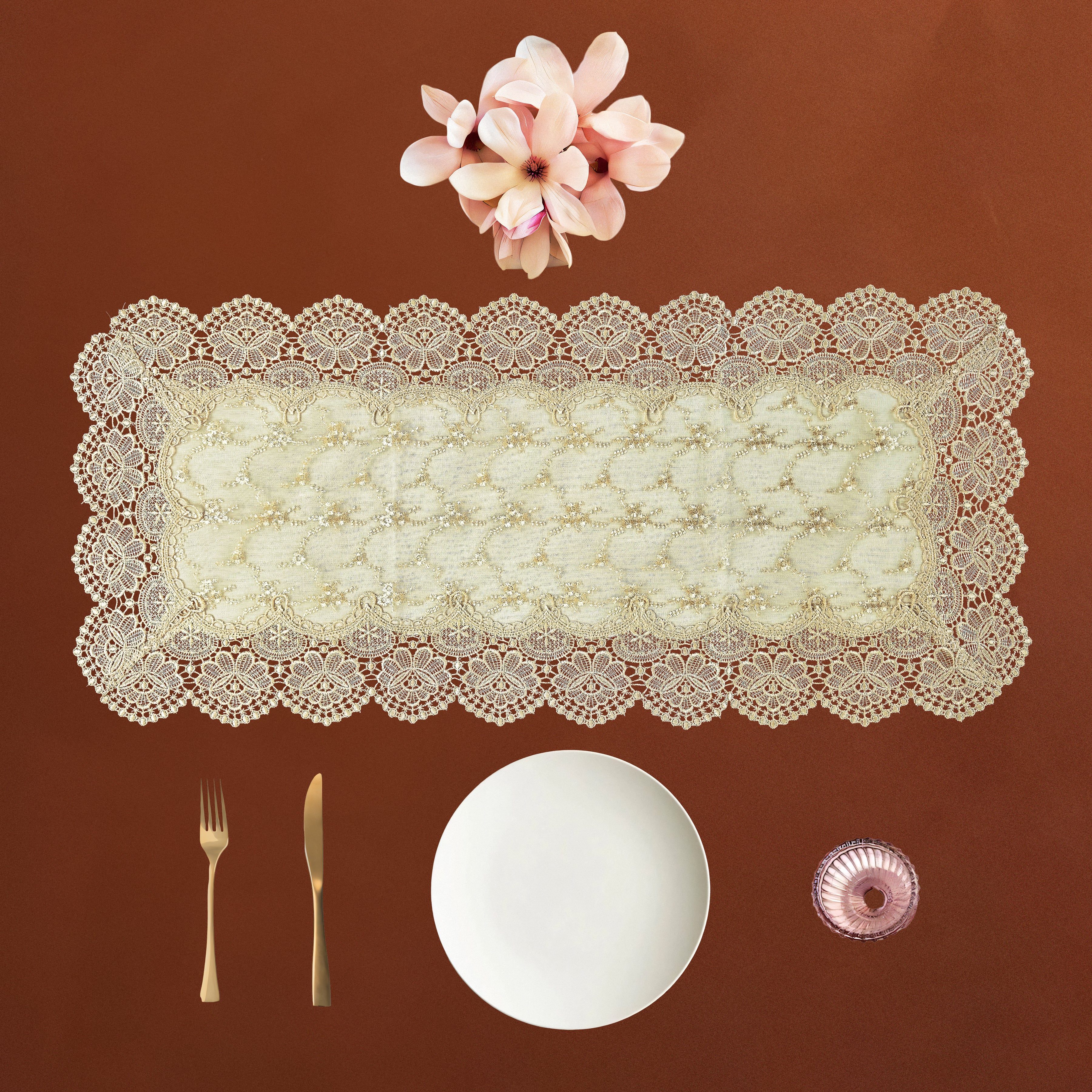 An off-white lace rectangular table runner with scalloped edges is displayed on a warm brown background, styled with a white plate, gold-toned cutlery, a small pink bowl, and pink flowers above it.