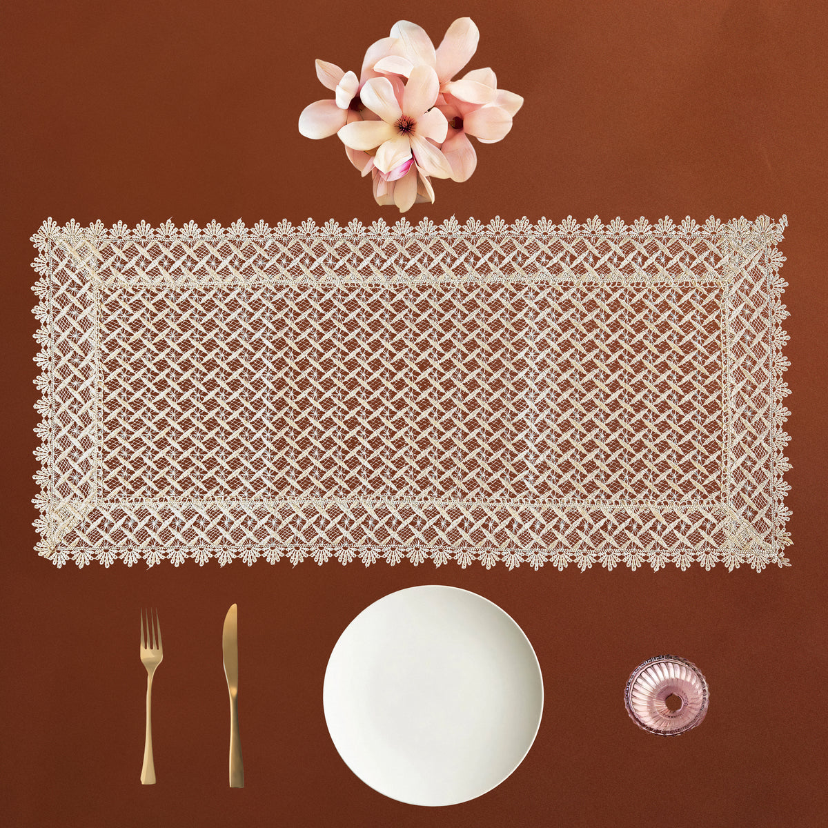 A rectangular white lace table runner with floral borders is displayed on a warm brown background, styled with a white plate, gold-toned fork and knife, and pink flowers above it.