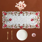 A rectangular embroidered table runner with a white damask center and a floral border in red, pink, and brown, styled on a brown background with a white plate, gold fork and knife, a small pink bowl, and pink flowers above.