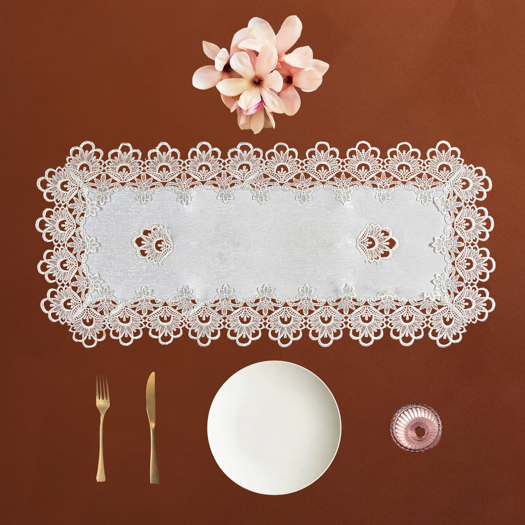 A rectangular white lace table runner with an intricate floral border, displayed on a warm brown background with a white plate, gold-toned fork and knife, and a small pink glass bowl.