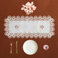 A rectangular white lace table runner with an intricate floral border, displayed on a warm brown background with a white plate, gold-toned fork and knife, and a small pink glass bowl.