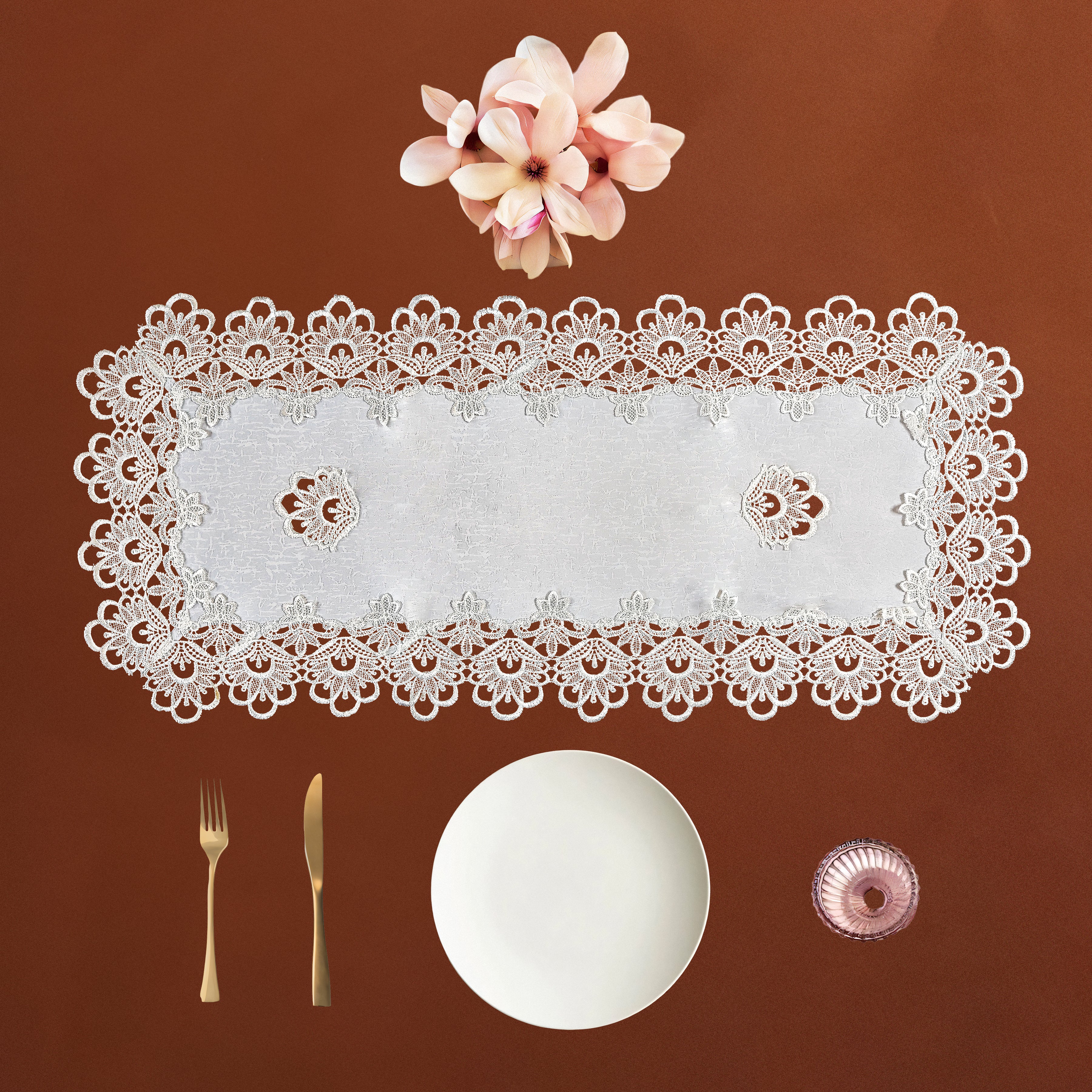 A rectangular white lace table runner with an intricate floral border, displayed on a warm brown background with a white plate, gold-toned fork and knife, and a small pink glass bowl.
