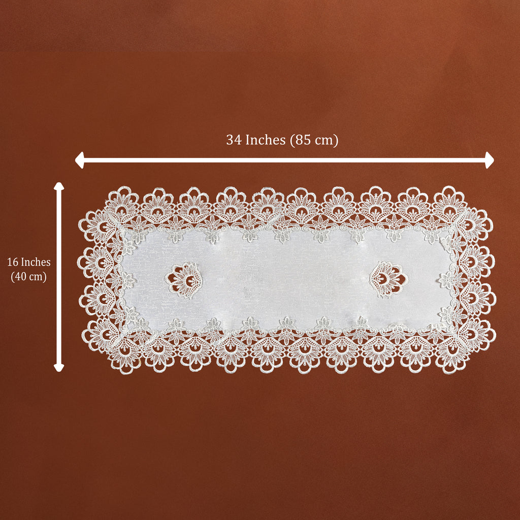 White rectangular lace doily with scalloped edges and two small floral cutouts, shown on a brown background with dimension arrows indicating 34 inches by 16 inches.
