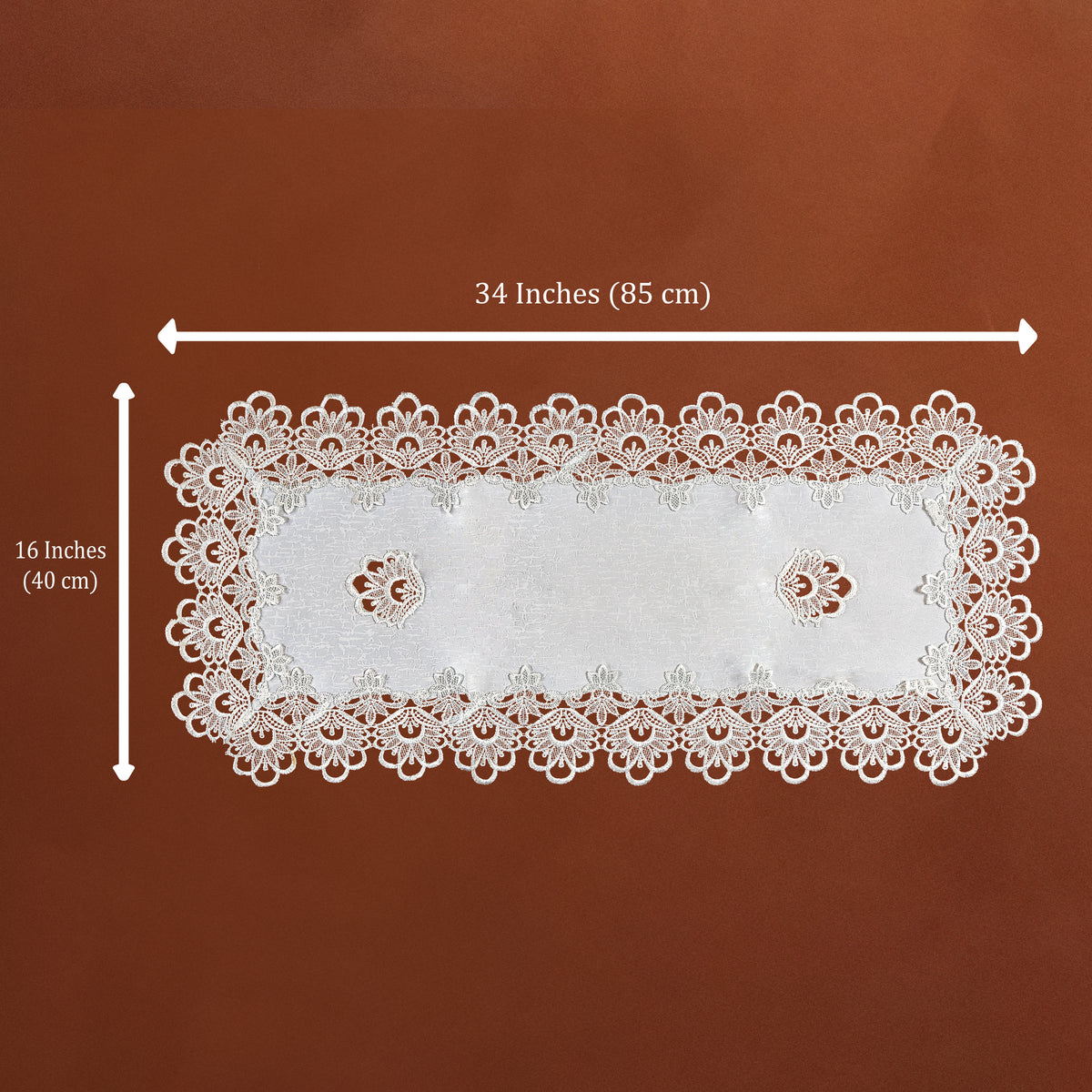 White rectangular lace doily with scalloped edges and two small floral cutouts, shown on a brown background with dimension arrows indicating 34 inches by 16 inches.