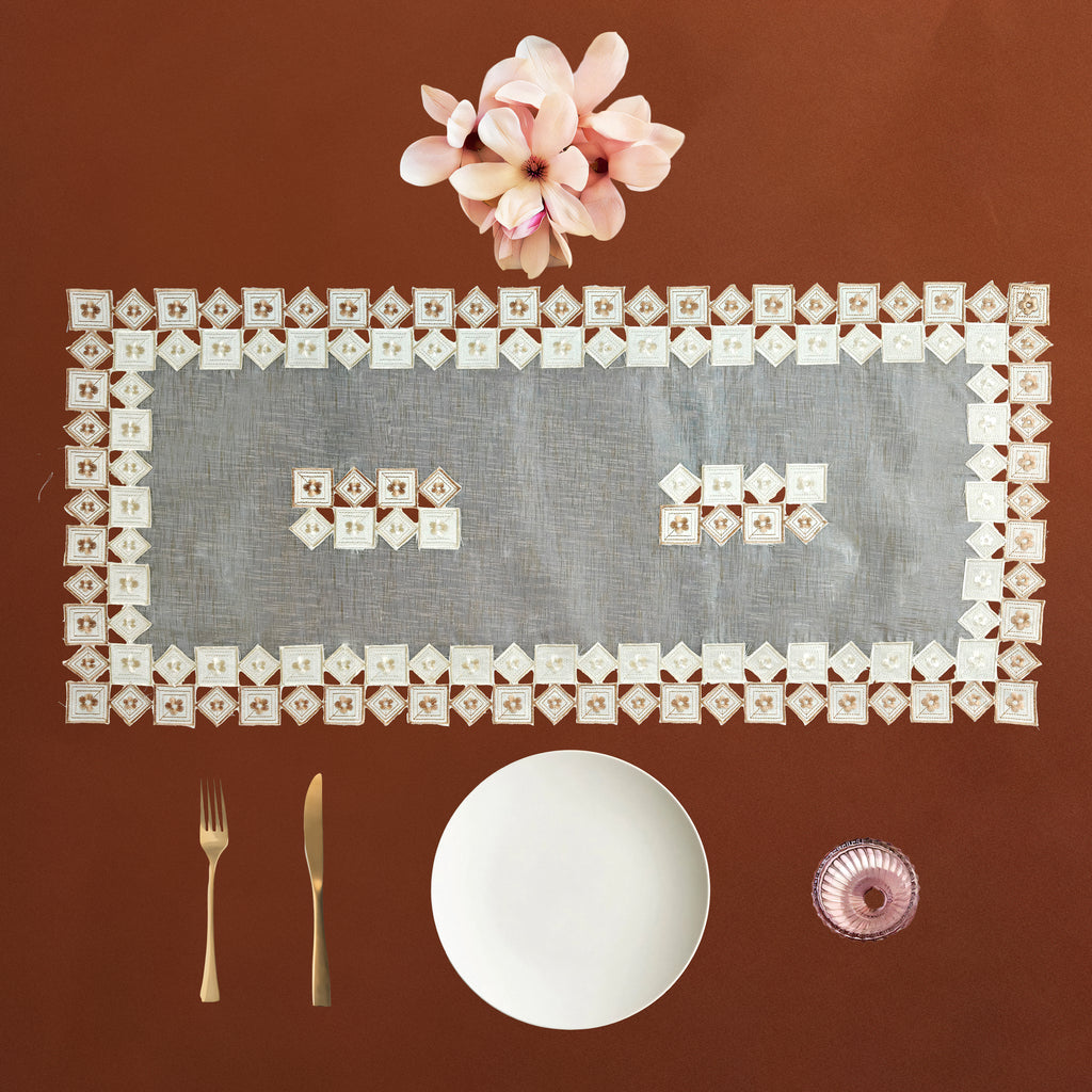 A square place setting flat lay on a warm brown background, featuring a clear rectangular table runner bordered with small white napkins, a white plate, gold-toned cutlery, a small pink glass, and a pink flower at the top.