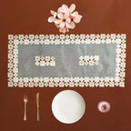A square place setting flat lay on a warm brown background, featuring a clear rectangular table runner bordered with small white napkins, a white plate, gold-toned cutlery, a small pink glass, and a pink flower at the top.