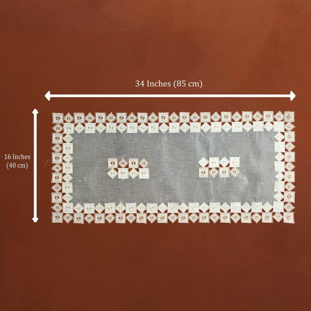A rectangular white lace textile with embroidered floral borders and two small embroidered motifs in the center, shown on a brown background with dimension arrows and text indicating 34 inches by 16 inches.