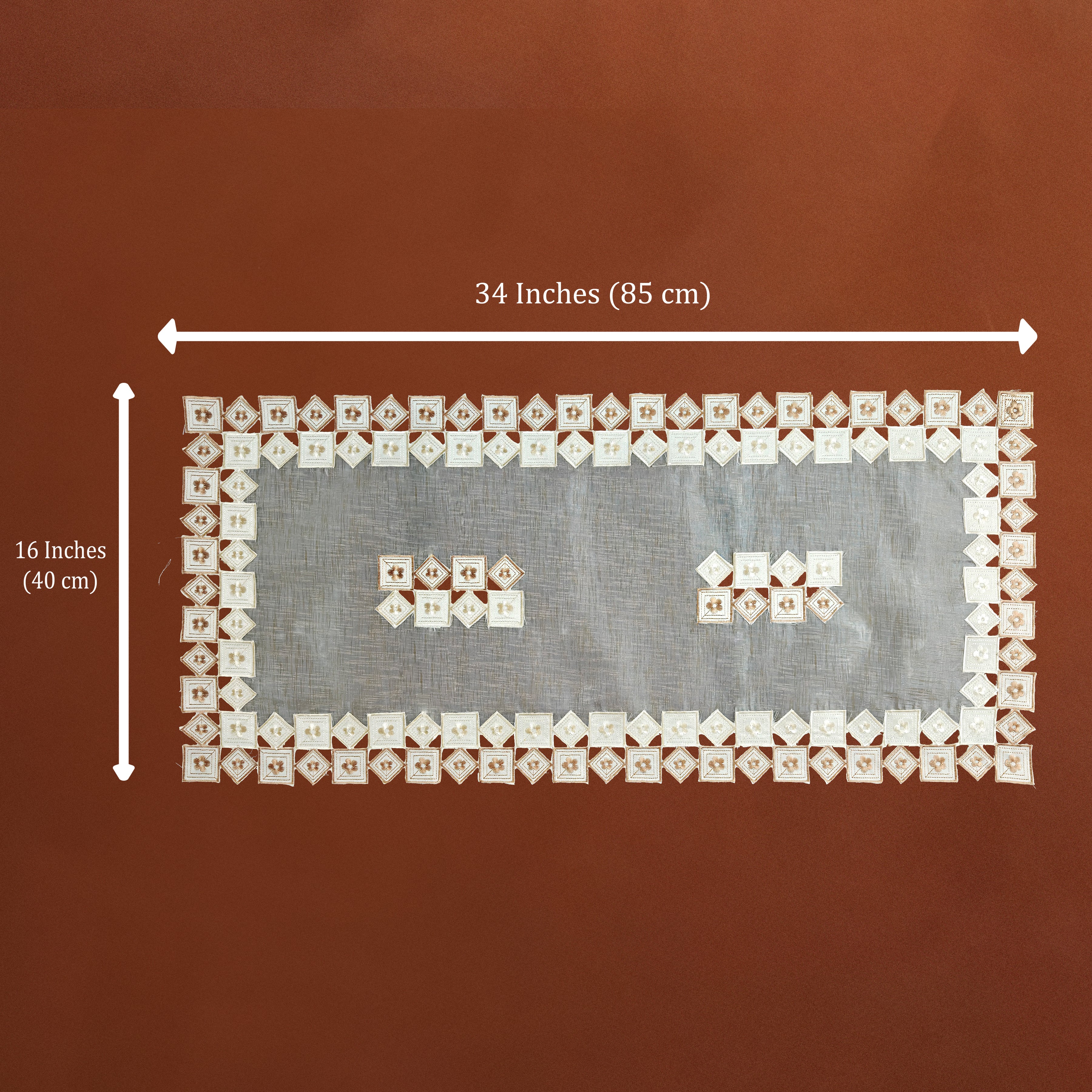 A rectangular white lace textile with embroidered floral borders and two small embroidered motifs in the center, shown on a brown background with dimension arrows and text indicating 34 inches by 16 inches.