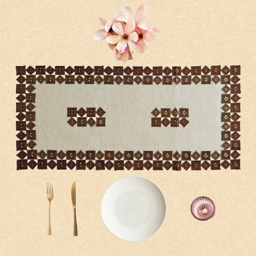 A decorative place setting on a beige textured background, featuring a rectangular placemat made of repeating dark brown diamond-shaped tiles, a white plate, gold-toned cutlery, a small pink glass bowl, and pink flowers at the top.