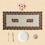 A decorative place setting on a beige textured background, featuring a rectangular placemat made of repeating dark brown diamond-shaped tiles, a white plate, gold-toned cutlery, a small pink glass bowl, and pink flowers at the top.